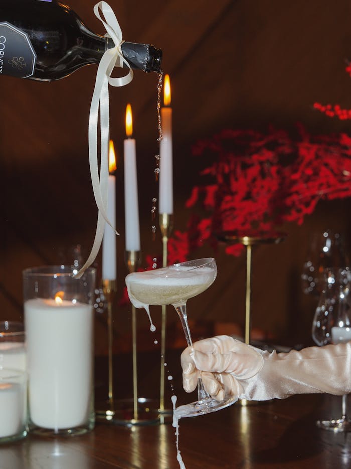 A sophisticated scene of champagne being poured into a glass amid warm candlelight and red floral decor.